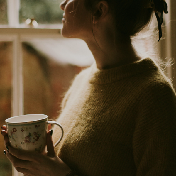 A woman with hands clasped around a cup of warm drink, looking satisfied and in balance, with a warm glow. 