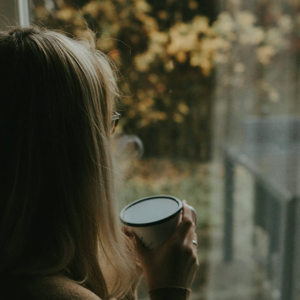 A woman looking out of the window to their garden, an autumn scene with a tree whose leaves have turned to autumn shades of yellow red and brwon. 