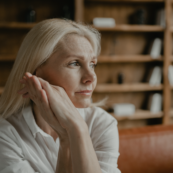 A woman looking pensive and off to the side in a shot where she is sat on a sofa in front of a bookshelf