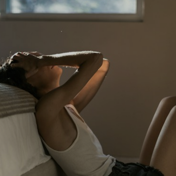 A woman sat on the floor leaning against the bed looking frustrated with her head in her hands
